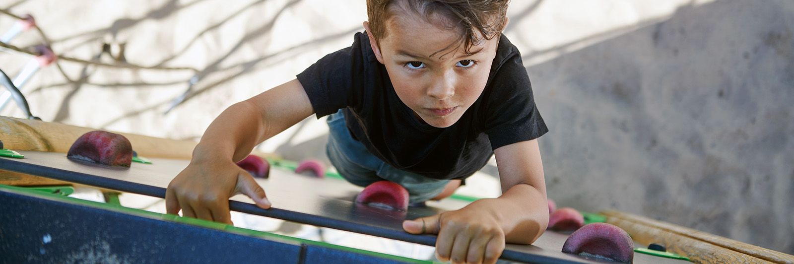 From above we see a boy climbing to the top on a climbing wall, he is looking upwards towards the camera planning his next move.